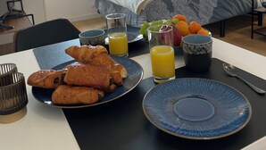 A neatly arranged breakfast table with croissants, fruit, and juice in a modern hotel room with a bed and a view of greenery. - The studio with its large, unoverlooked terrace, in a quiet location with private parking (Saint-Estève)