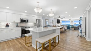 A modern kitchen with white cabinetry, a central island with bar stools, stainless steel appliances, and pendant lighting. - Fall Deals + Snowbird Rates | Gulf Front Beach Escape (Panama City Beach)