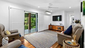 A modern living room with a large sliding glass door leading to a balcony, a flat-screen TV mounted on the wall, and a wooden entertainment unit. - Family Friendly Walk to Beach Tropical Retreat + BBQ Deck (Point Vernon)