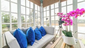 Un solárium con un ventanal grande, un sofá gris con cojines azules y una orquídea en maceta.