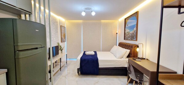 A compact hotel room with a bed, bedside table, lamp, and a refrigerator.