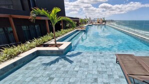 A long, narrow swimming pool with clear blue water, surrounded by tiled flooring and palm trees. - COARM0106 - Edificio Art Studio (Salvador)