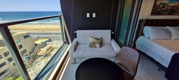 Un balcón con vista a la playa y al océano. Hay un sofá gris con un cojín estampado y una silla marrón. Se ve una cama con ropa blanca a la derecha.