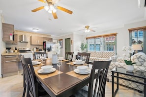 A modern dining area with a wooden table set for four, surrounded by chairs. There are ceiling fans above, and the room has a large window with a view of buildings outside. - Charming 2BR Home • Outdoor Kitchen (Noord)