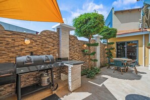 An outdoor patio with a barbecue grill, a dining table with chairs, and a small tree.
