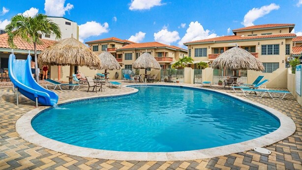 A swimming pool with lounge chairs and a slide, surrounded by buildings with red roofs and thatched umbrellas.