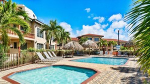 A resort with two swimming pools, lounge chairs, palm trees, and a building with a red roof.