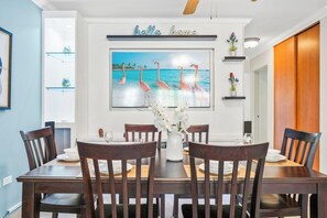A dining area with a table set for six, surrounded by wooden chairs, a white vase with flowers, and a wall-mounted shelf with decorative items.