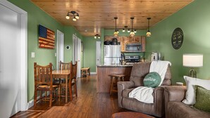 A cozy living room with a brown leather sofa, wooden dining table, and chairs. A kitchen with wooden cabinets and a stainless steel refrigerator is visible in the background. - Ozark Mountain Lake Home (Branson West)