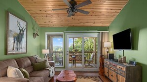 A living room with a wooden ceiling fan, a leather couch, a coffee table, a TV, and a sliding glass door leading to an outdoor patio. - Ozark Mountain Lake Home (Branson West)