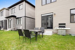 A modern two-story house with a patio area featuring a dining table and chairs, a grill, and a small shed.