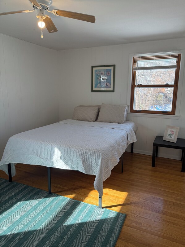 Un dormitorio con una cama, ventilador de techo, ventana con persianas y un cuadro en la pared.