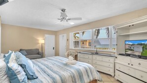 Un dormitorio con cama, ventilador de techo, cómoda, televisor y vista a una casa desde la ventana.
