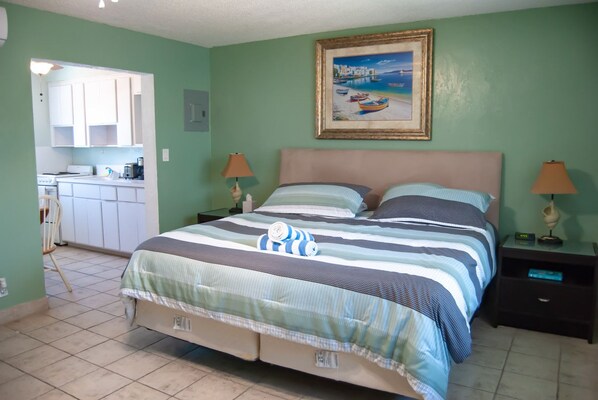 Quarto com cama, duas mesas de cabeceira, um criado-mudo, um quadro e uma cozinha visível por uma porta aberta.