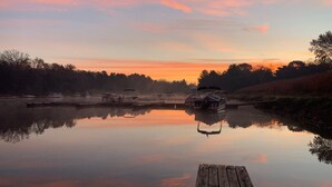 A calm river at sunset with a wooden dock extending into the water. Boats are docked along the riverbank.