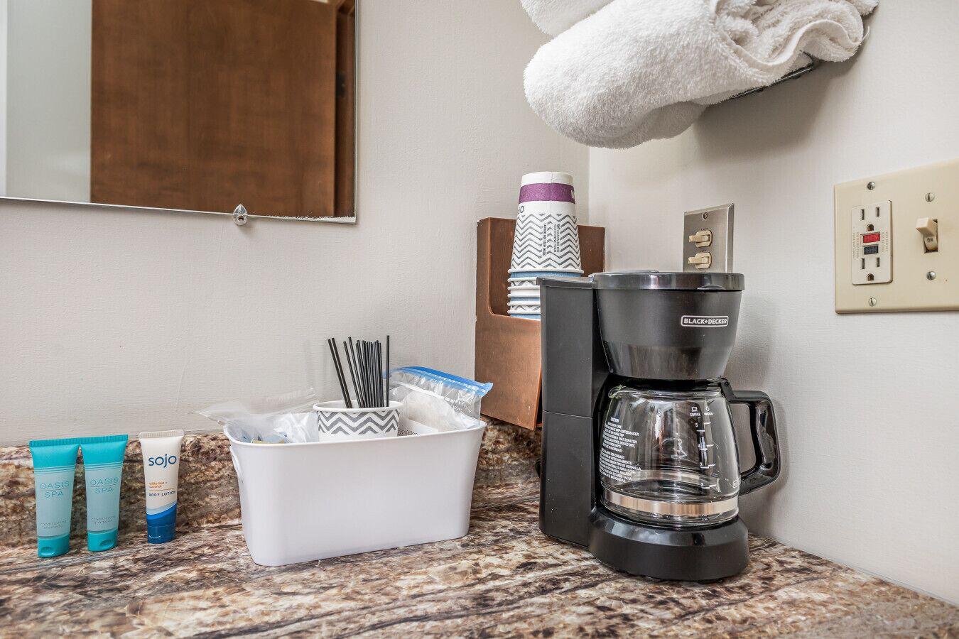 Una encimera de baño con cafetera, artículos de aseo y un perchero para toallas.