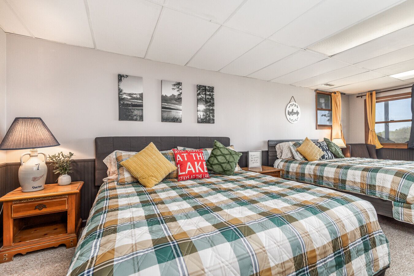 Quarto de hotel com duas camas, mesa de cabeceira com abajur, placa de boas-vindas emoldurada e três quadros pendurados na parede.