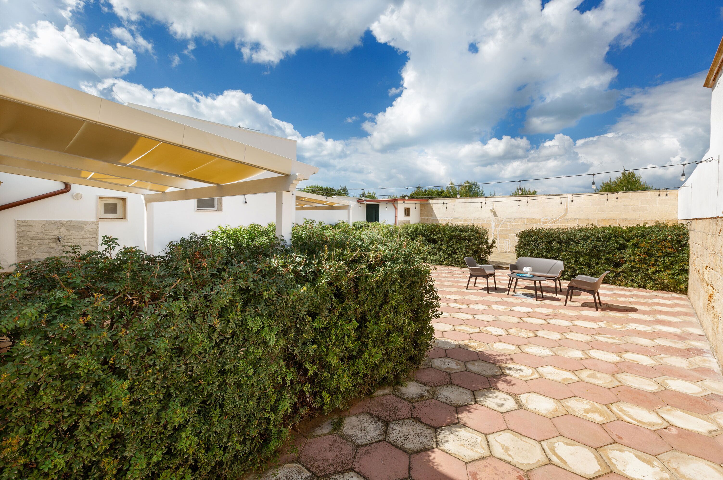 A covered patio area with hexagonal tiles, a seating arrangement, and a hedge.