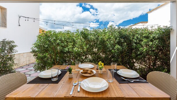 Mesa de madera dispuesta para dos personas, con platos, cubiertos y vasos, bajo una pérgola con vistas a un seto verde exuberante y un cielo parcialmente nublado.