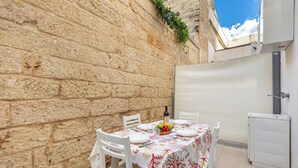 Un área de comedor al aire libre con paredes de piedra, una mesa lista para dos personas, sillas, un frutero pequeño y un armario blanco.