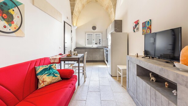 Una cocina moderna con horno empotrado, un comedor con sofá rojo y una pequeña mesa, y un televisor sobre un mueble de madera.