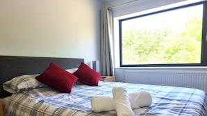 A neatly made bed with plaid bedding and two red pillows, a window with a view of trees, and a radiator under the window. - Whole house with parking and garden in Conwy. Pets welcome (Conwy)