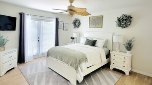 Un dormitorio con cama, mesitas de noche, ventilador de techo, ventana con persianas y un adorno en la pared.