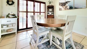Un comedor con mesa de madera, sillas blancas, una araña, una estantería y vistas a través de un ventanal.