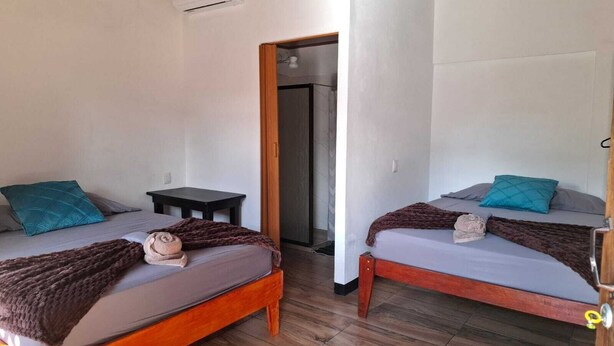 Une chambre avec deux lits en bois, chacun doté d’un matelas gris et d’oreillers turquoise. Une petite table en bois se trouve entre les lits, et un appareil de climatisation est fixé au mur.