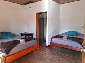 Ένα δωμάτιο με δύο ξύλινα κρεβάτια, το καθένα με ένα γκρι στρώμα και γαλαζοπράσινες μαξιλάρες. Ανάμεσα στα κρεβάτια υπάρχει ένα μικρό ξύλινο τραπέζι και στον τοίχο είναι τοποθετημένη μια μονάδα κλιματισμού.