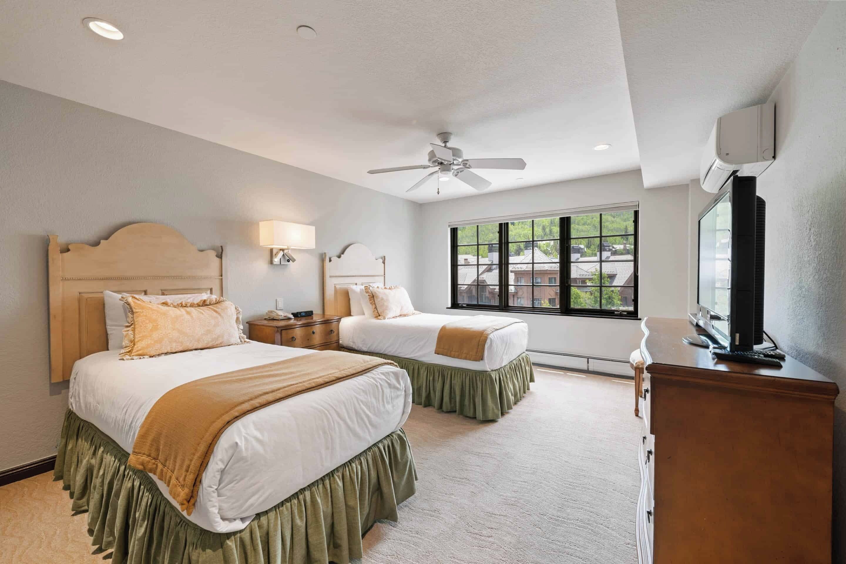 A hotel room with two beds, a ceiling fan, a window with a view, and a TV on a dresser.