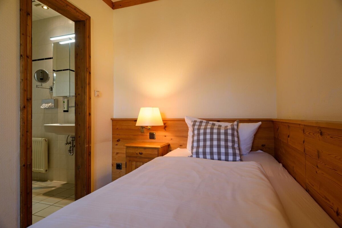 Ein Hotelzimmer mit Bett, Nachttisch mit Lampe, Holz-Kopfschild, Badezimmer mit Spiegel und Heizkörper.