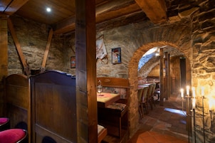 A rustic interior with stone walls, wooden beams, a dining area with tables and chairs, and a small alcove with a bed.