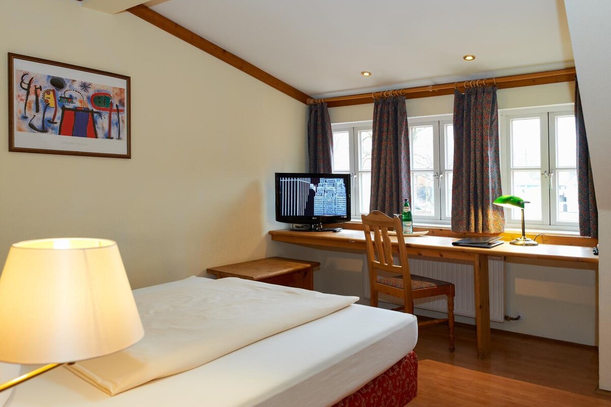 Ein Hotelzimmer mit Bett, Schreibtisch, Computer, Stuhl, Lampe und einem Vorhangfenster.