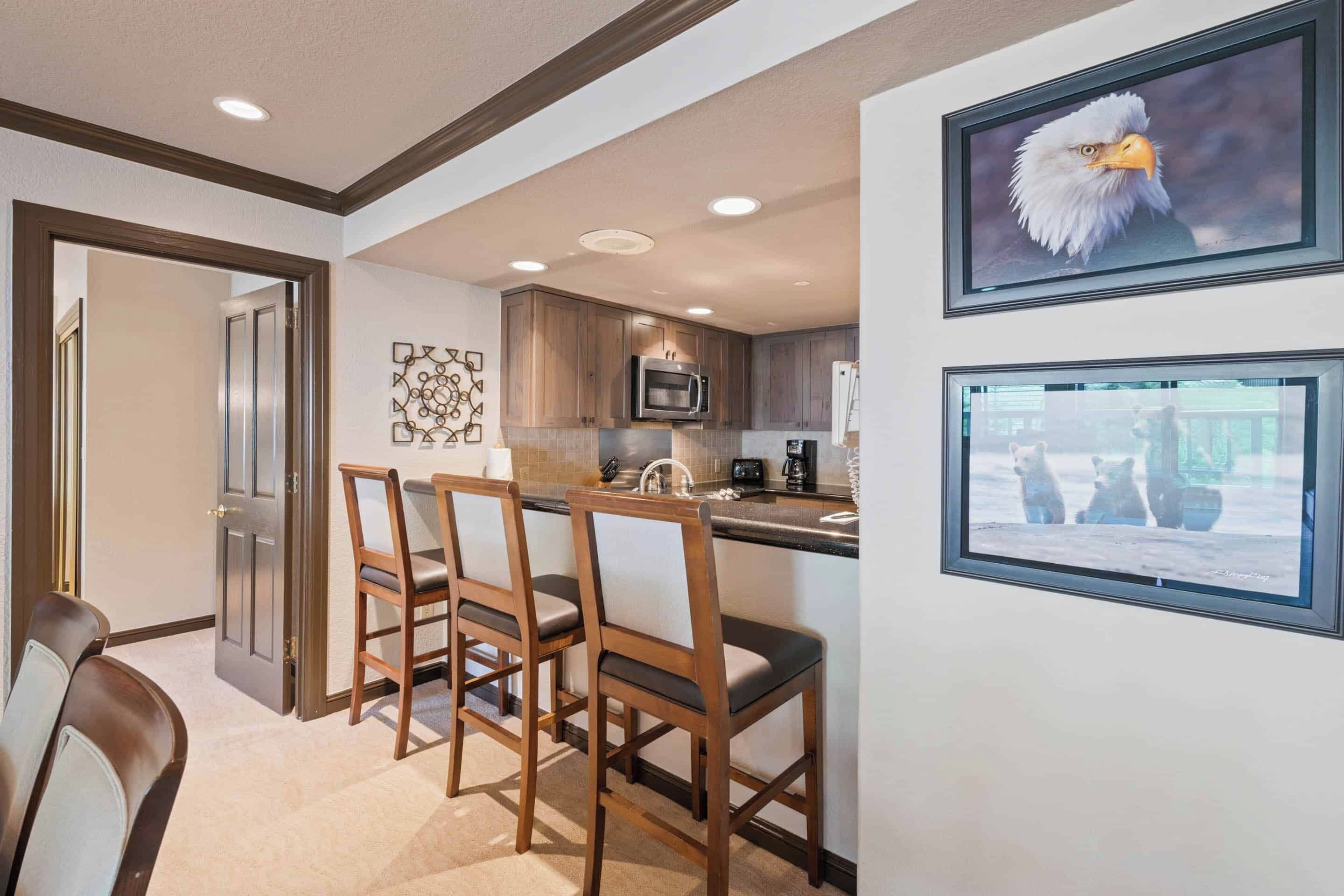 A modern kitchen with wooden cabinets, a microwave, and a coffee maker. There are three wooden chairs and a dining table.