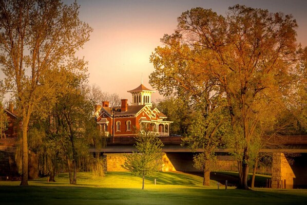 A large, historic building with a prominent tower and multiple chimneys, surrounded by lush trees and a well-maintained lawn.