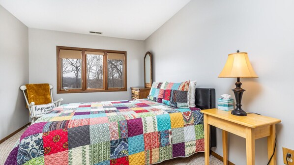 A bedroom with a colorful quilted bedspread, a wooden nightstand with a lamp, a chair with a 'happiness is home' sign, and a window with a view of bare trees.
