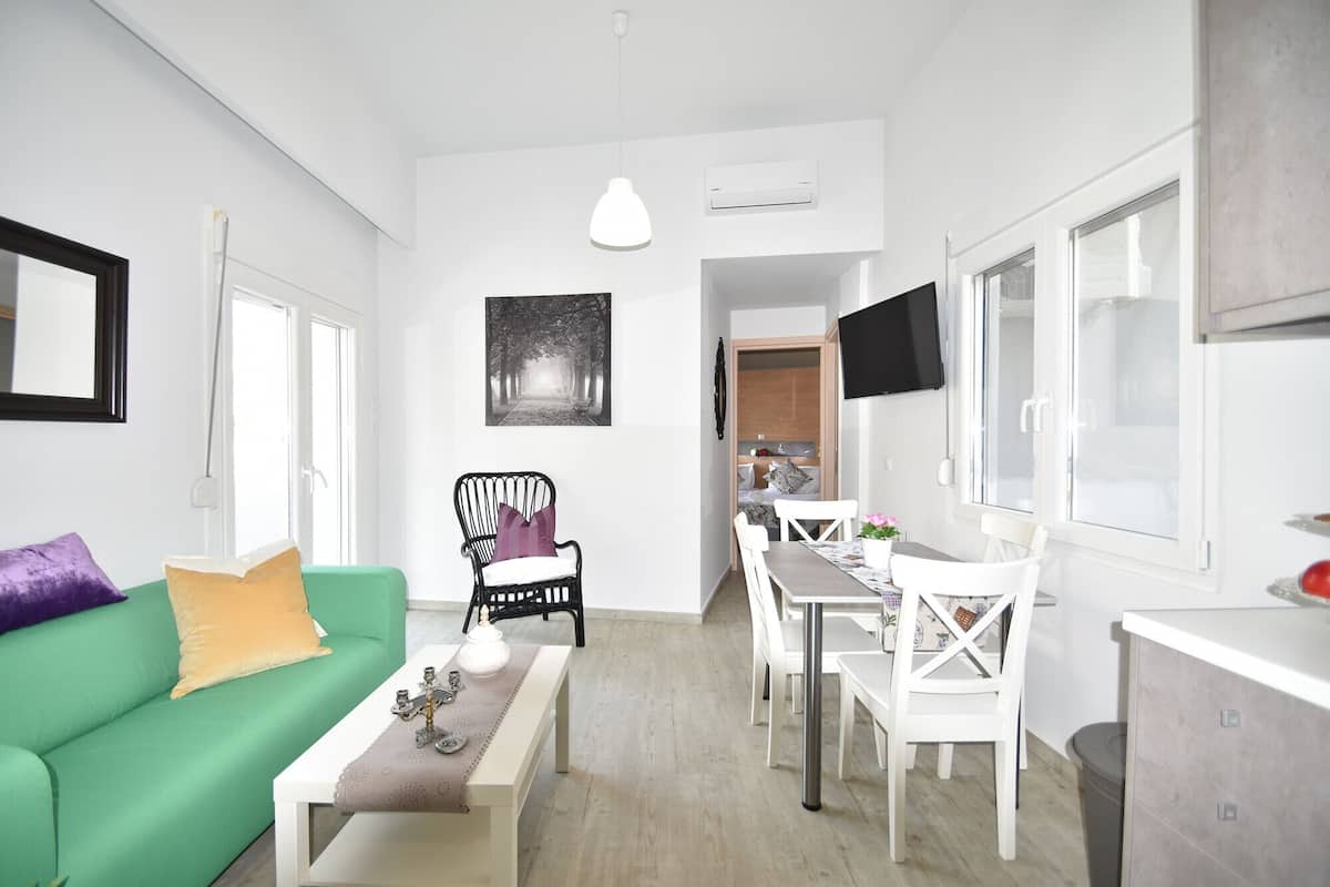 Ein modernes Wohnzimmer mit einem grünen Sofa, einem Esstisch mit weißen Stühlen, einem schwarzen Sessel mit weißem Kissen und einem flachbildfernseher, der an der Wand montiert ist.