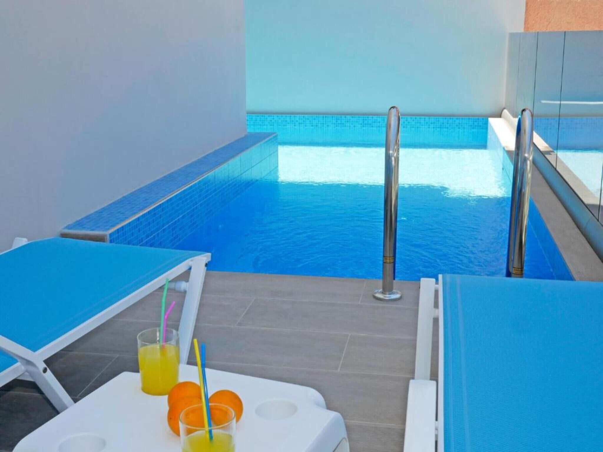 A poolside area with a blue-tiled pool, lounge chairs, and a table with drinks and fruit.
