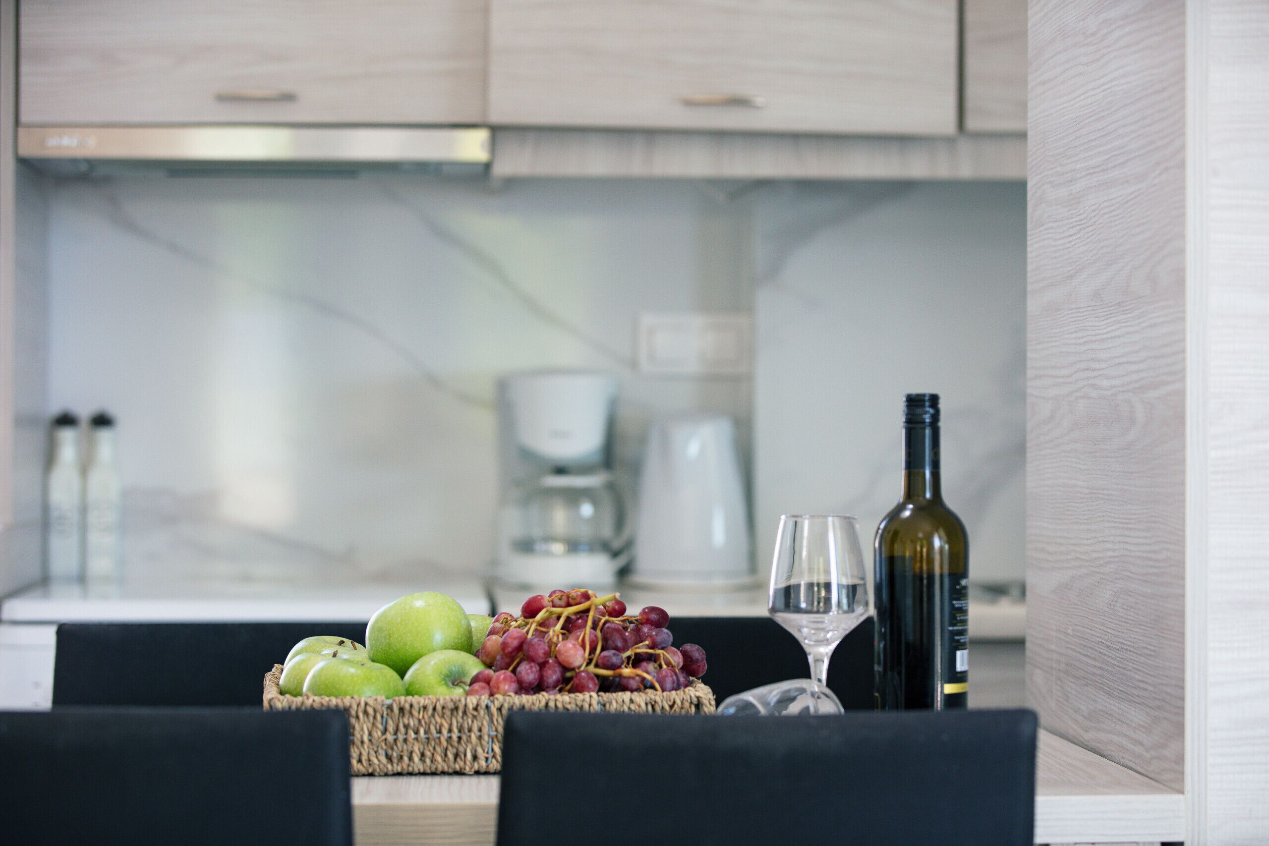 Una encimera de cocina con una cesta de manzanas verdes y uvas rojas, una botella de vino y dos copas de vino.