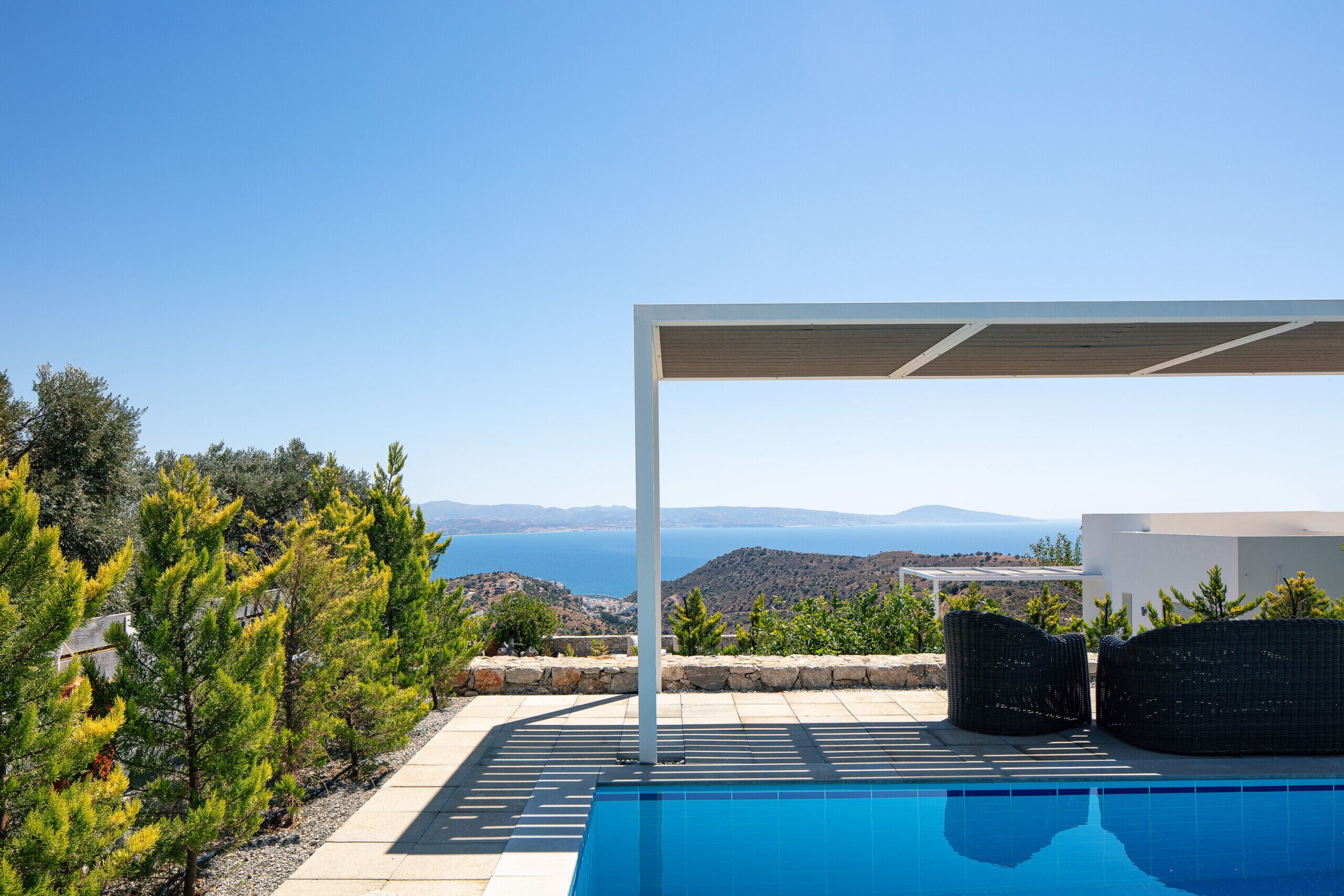 A poolside area with a pergola, outdoor furniture, and a view of the sea and hills.