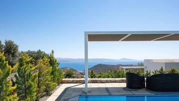 A poolside area with a pergola, outdoor furniture, and a view of the sea and hills.