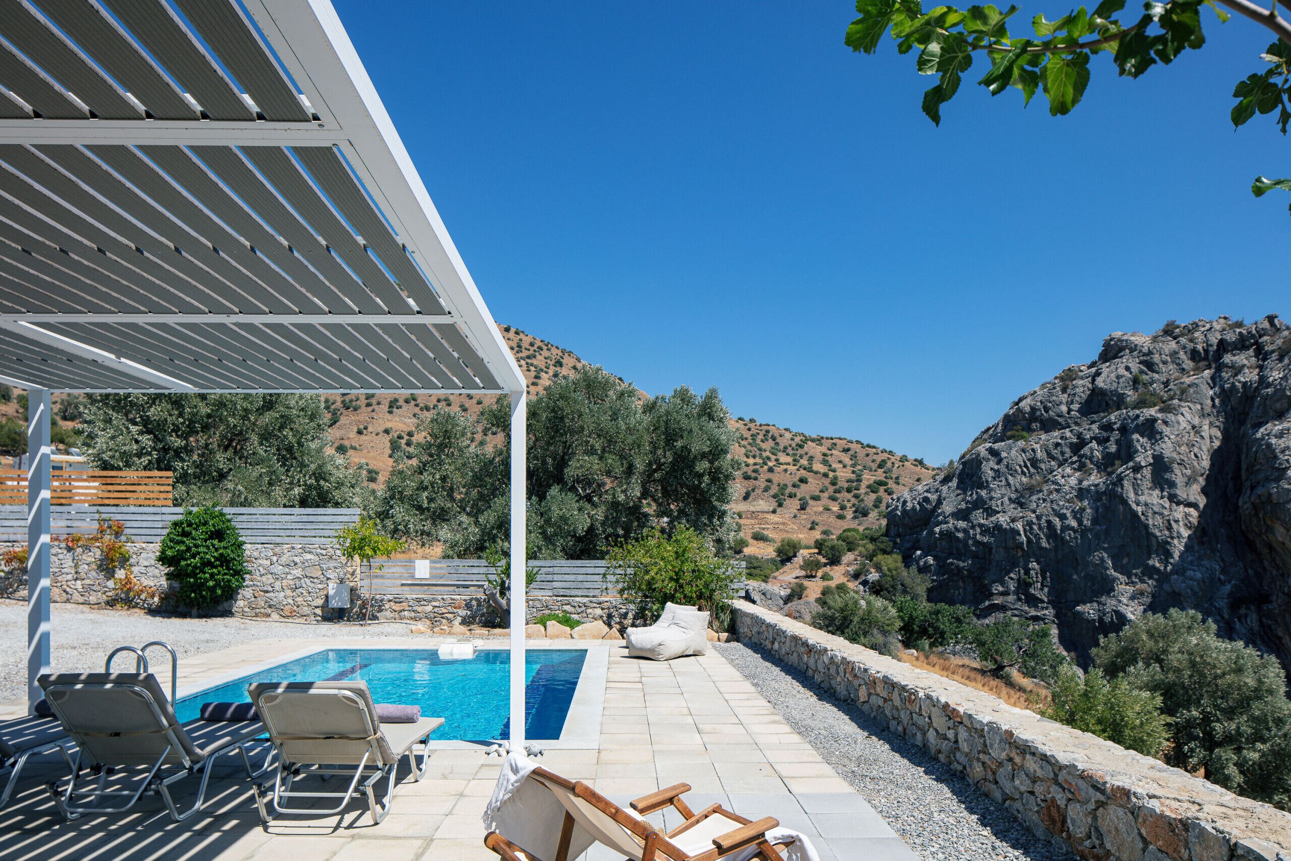 A pool area with lounge chairs, a pergola, and a view of rocky hills.