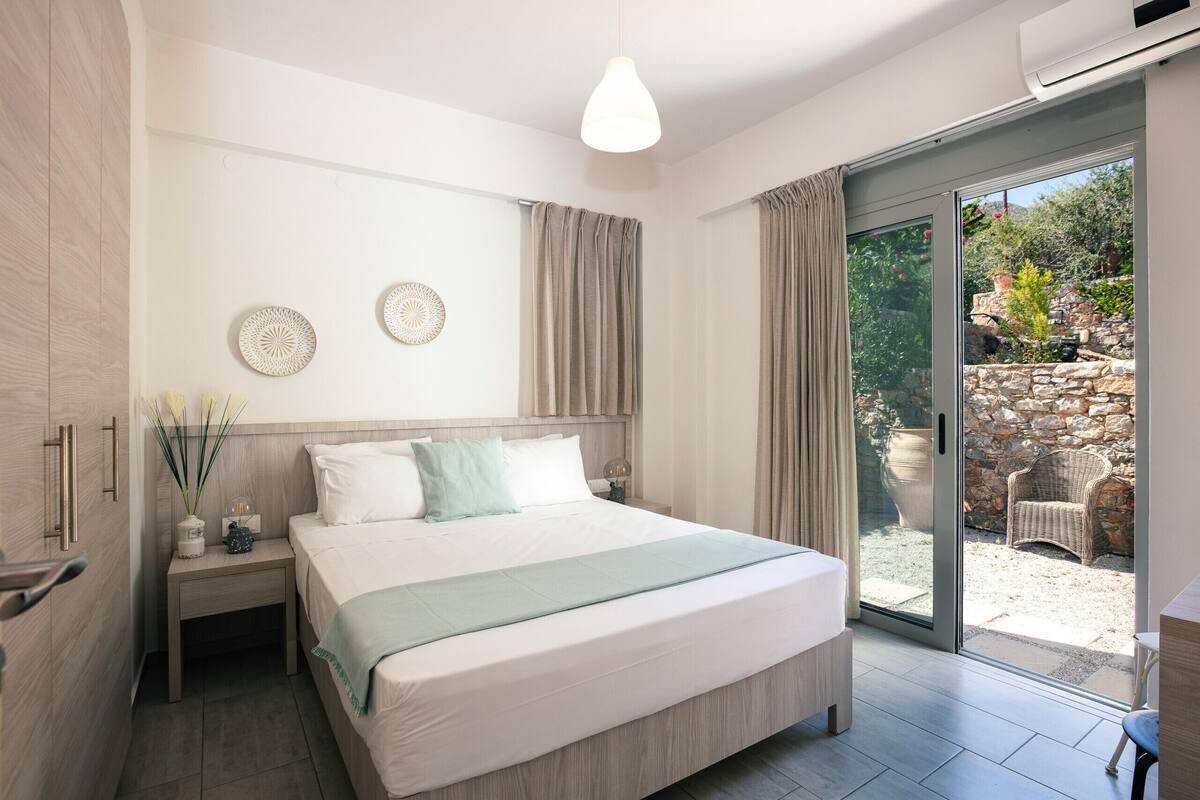 A modern bedroom with a large bed, a nightstand, a wall-mounted lamp, and a sliding glass door leading to an outdoor area.