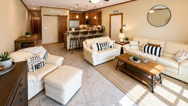 A modern living room with a sofa, ottoman, and coffee table, a kitchen area, and a ceiling fan. - Paraiso Del Rio | 3BDRM | Guadalupe River (New Braunfels)