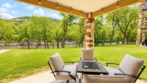 An outdoor patio with a table and chairs, a stone pillar, and a view of a grassy area with trees and a body of water. - Paraiso Del Rio | 3BDRM | Guadalupe River (New Braunfels)