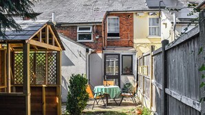Un jardin arrière d’une maison, aménagé avec une cabane en bois pour les enfants, une petite table avec des chaises et un espace potager.