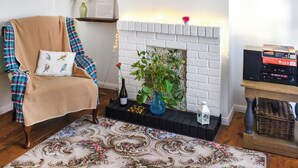 Un salon avec une cheminée en briques blanches, un fauteuil à carreaux, un tapis à motifs floraux, une petite table avec un vase et une bouteille, et un haut-parleur posé sur un support.