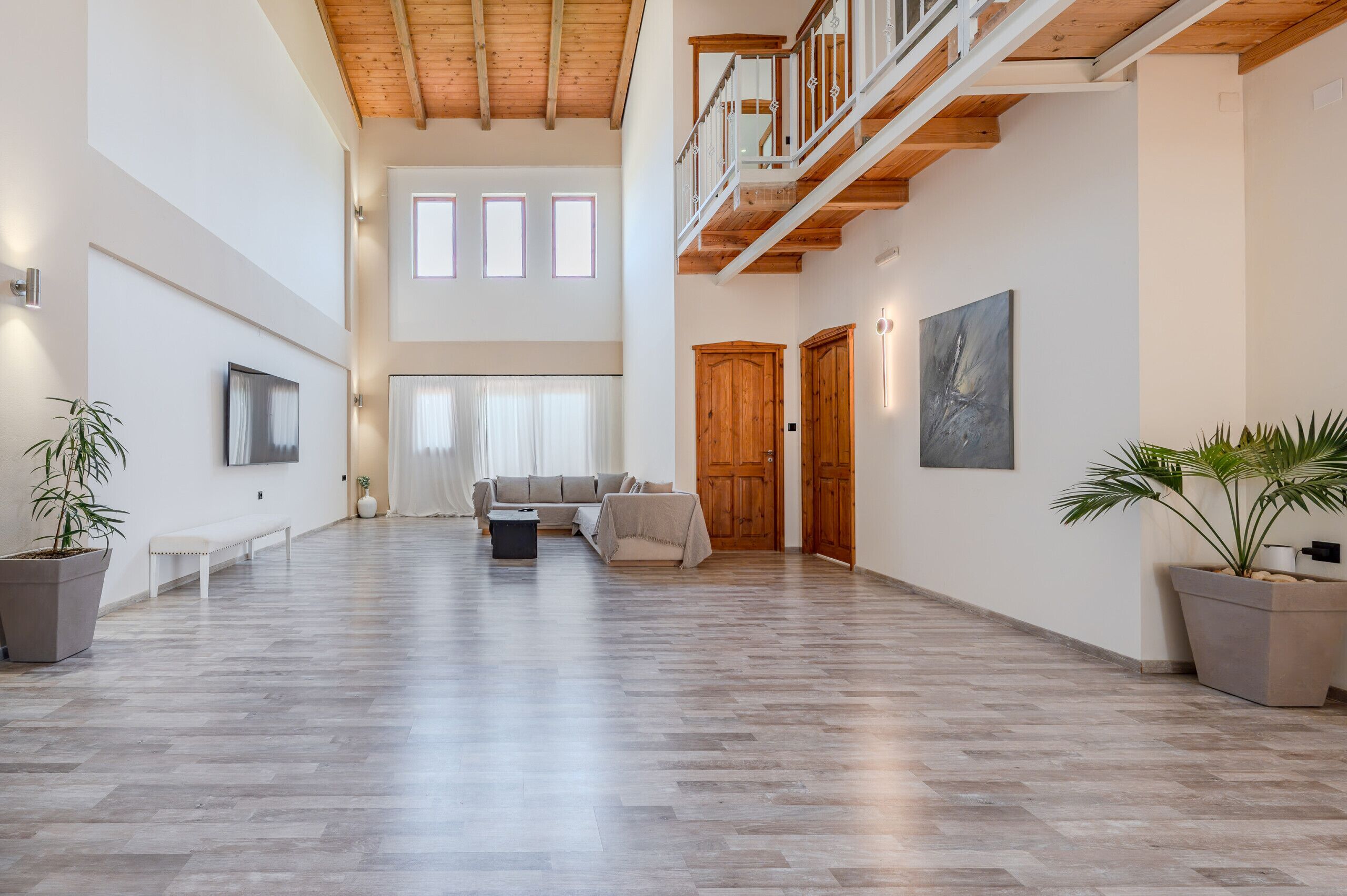 Een ruime woonkamer met een hoge plafond, houten balken, een bank, een bijzettafeltje en potplanten.