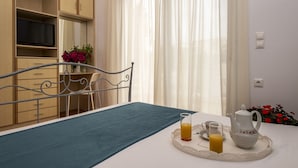 Ein ordentlich eingerichtetes Schlafzimmer mit einem Bett, einem Nachttisch mit Teekanne und Gläsern, einem Fernseher auf einem Regal und einem Fenster mit durchscheinenden Vorhängen.